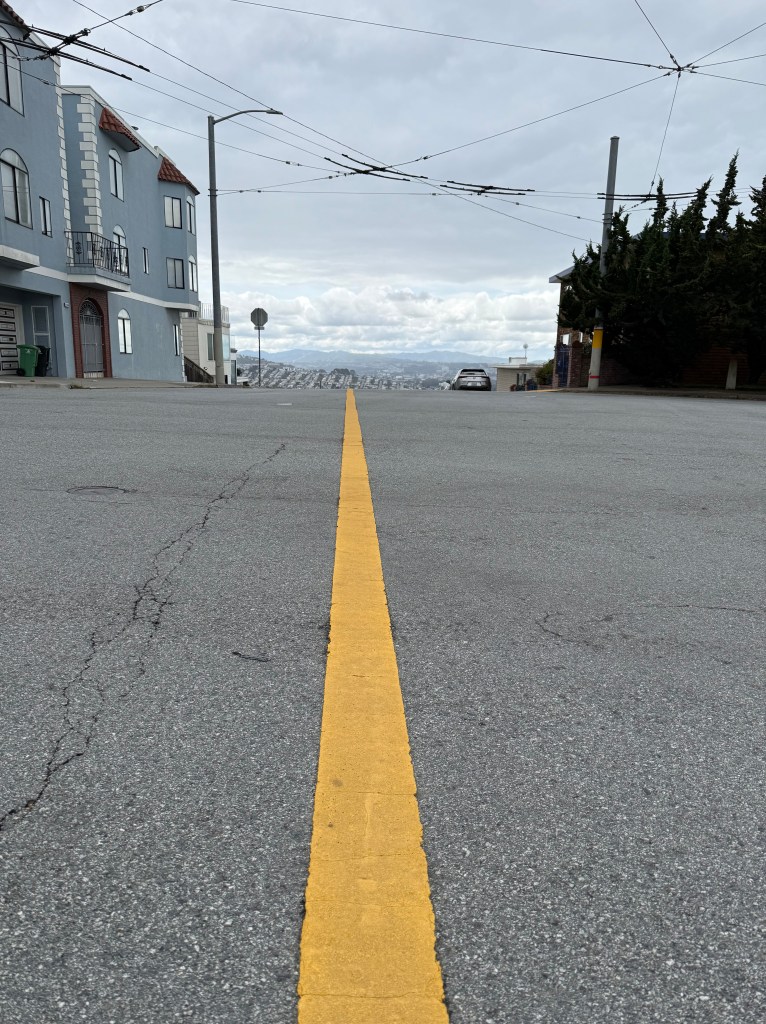 A line in the street pointing to Daly City and the San Bruno Mountain Ridge to the south. The faint ridge in the background was where Sergeant Ortega of the Spanish Army scaled where current Sharp Park Road goes over to Skyline College, and was the first European discovery to realize there was a huge inland bay that could connect to the Golden Gate by the Presidio.
